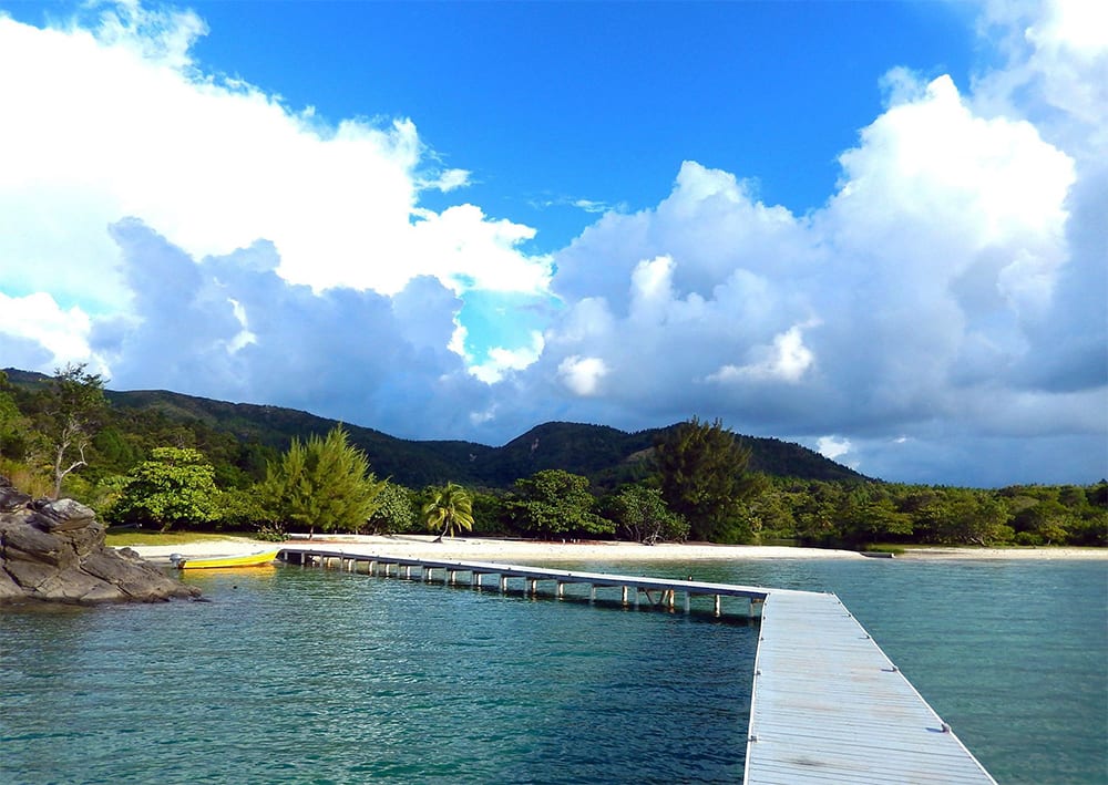 white jetty across blue sea to a sandy beach with bushes and trees behind and blue skies