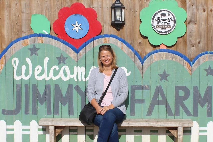 Sally from The Foodie Travel Guide sitting in front of a Welcome to Jimmy's Farm sign wearing a lavender cardigan and jeans