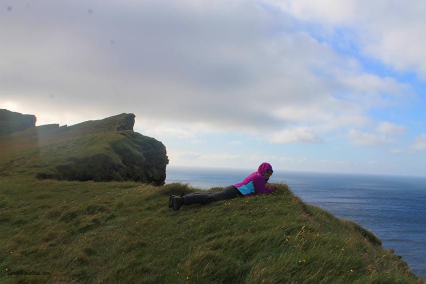 Woman wearing a pink and blue hoodie lying on a grass cliff looking out over the sea