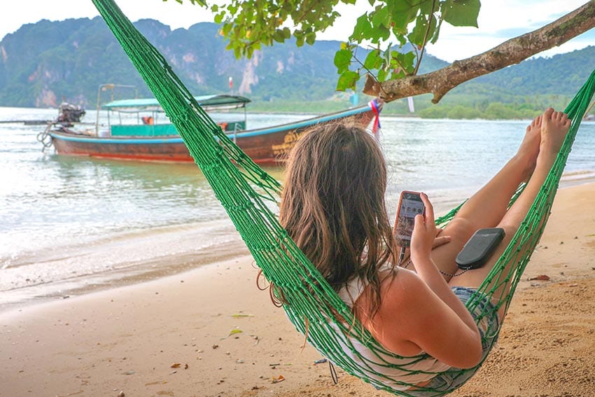 Girl sitting in a green hammock with her back to the camera looking at her mobile phone with a Thai boat in the background on the sea
