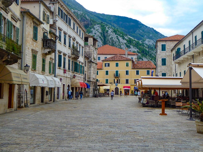 Old Town Kotor showing a cobbled street restaurants with awnings on the right and shops with awnings on the left. A yellow building in the distance with hills behind. 