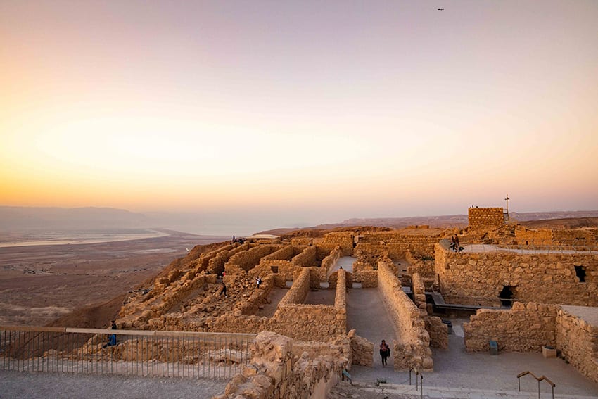 the ruins of Masada Fortress with the sun rising in the background. The walls show the rectangular layout of the rooms in the fort.