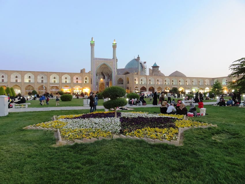 Naqs-e Jahan Square with arched sand coloured buildings in the background with three domes on the right and a larger arched entrance in the middle. In the foreground is a grassy area with a seven sided star planted with white, purple and yellow flowers and a tree in the middle. People are sitting on the grass or walking in the grounds.