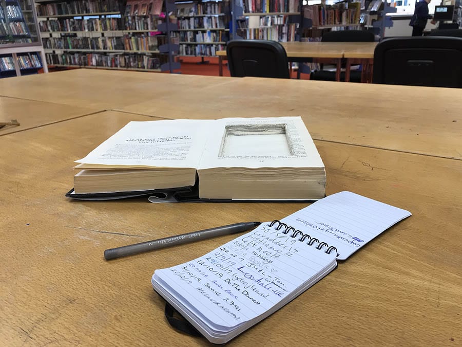 open book with a portion cut out with a note pad and pen beside it. In the background are library shelves full of books and chairs and tables
