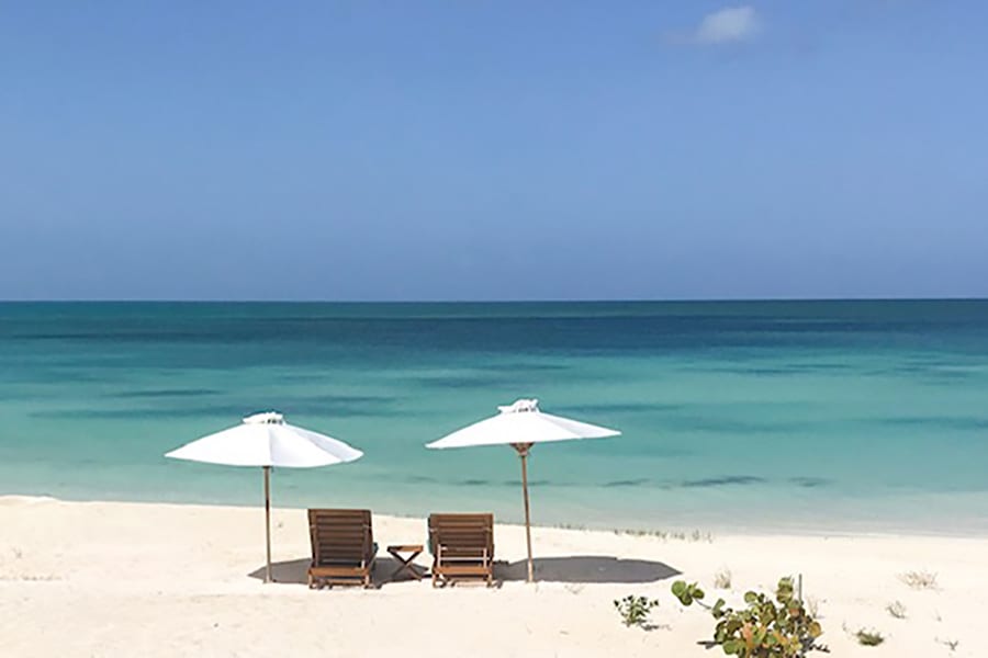 two sun loungers under white parasols on a white and pink sand beach next to the turquoise and blue sea with a clear blue sky above