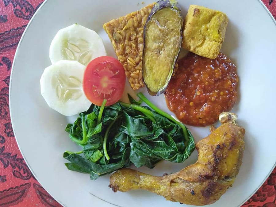 Ayam Goreng - Balinese Fried Chicken on a white plate on red tablecloth with fried chicken leg, greens, sliced tomato and cucumber, tempe, fried aubergine, corn fritter and tomato sambal
