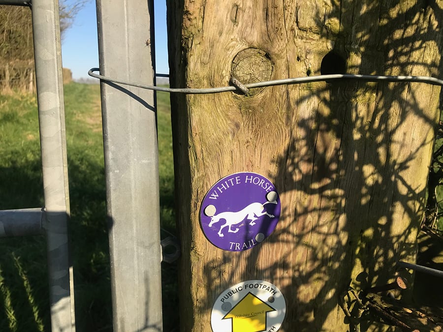 purple disk with White Horse Trail writing in white and and a white horse above a white Public Footpath disk with yellow up arrow saying Wiltshire Council