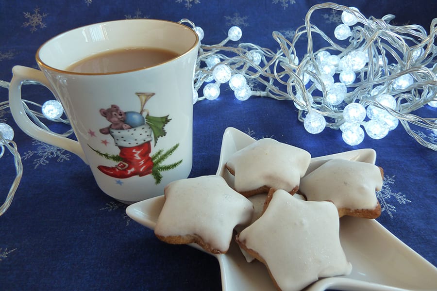 tradition star shaped Lebkuchen biscuits with icing sugar topping next to a cup of tea in a mug with a red Christmas boot on it and toys coming out the top all sitting on a blue fabric with silver snowflakes and lit by some white fairy lights