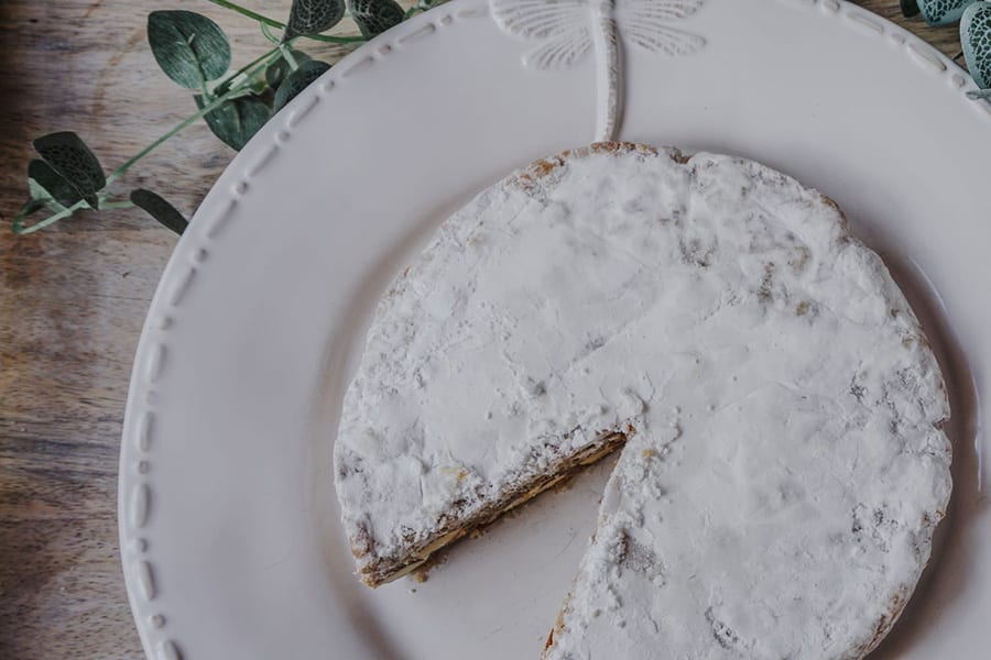 round cake topped with icing sugar sitting on a round white plate with raised dragonfly motif. In the background is some ivy.