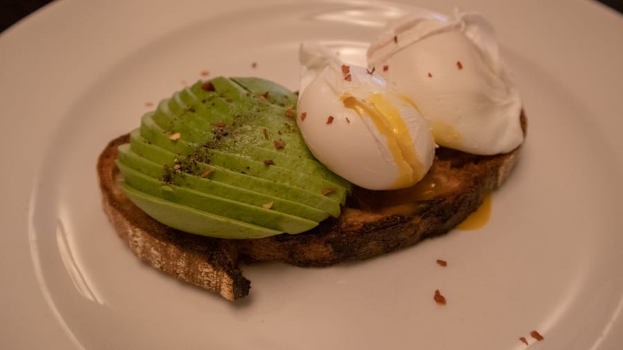 sourdough toast with sliced avocado and two poached eggs topped with pepper and chilli flakes. One of the eggs is split and the yellow yolk is oozing out