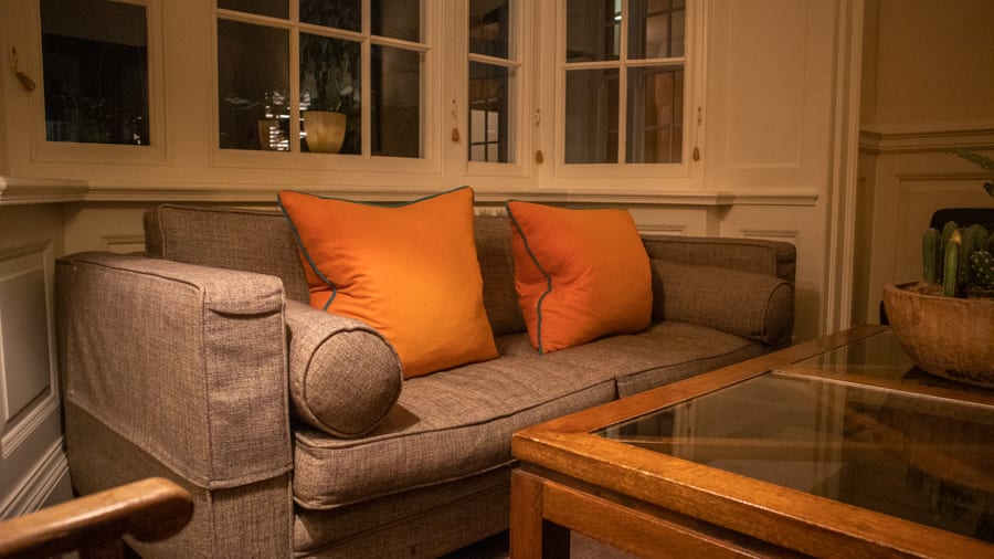 grey sofa with two orange square cushions piped in grey underneath windows with a glass topped table in front