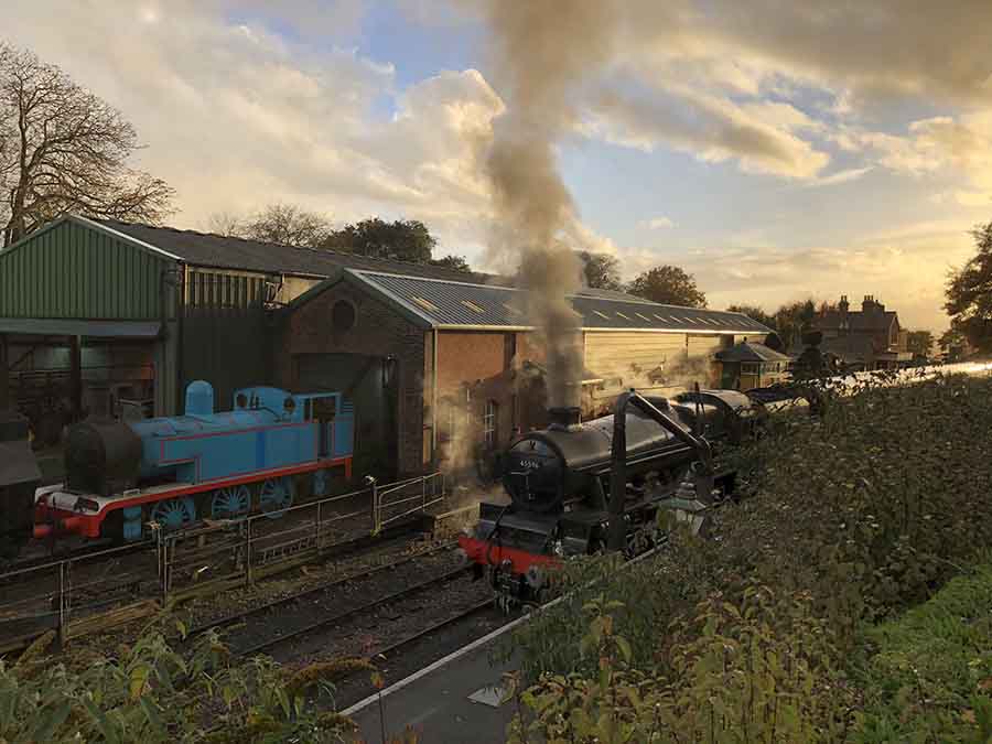 railway yard with a black steam train in front and a blue steam train coming out of a shed beside it