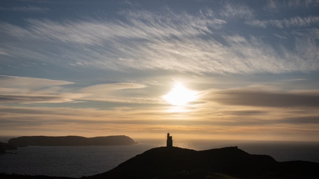 silhouette of a tower on at hill overlooking the sea with the setting sun and clouds in the sky