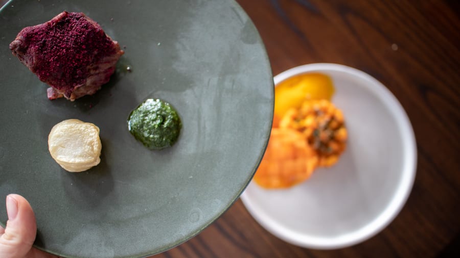 Beef with beetroot powder, wild garlic and potato stack on a gry plate with white plate below with carrot lattice, carrot puree