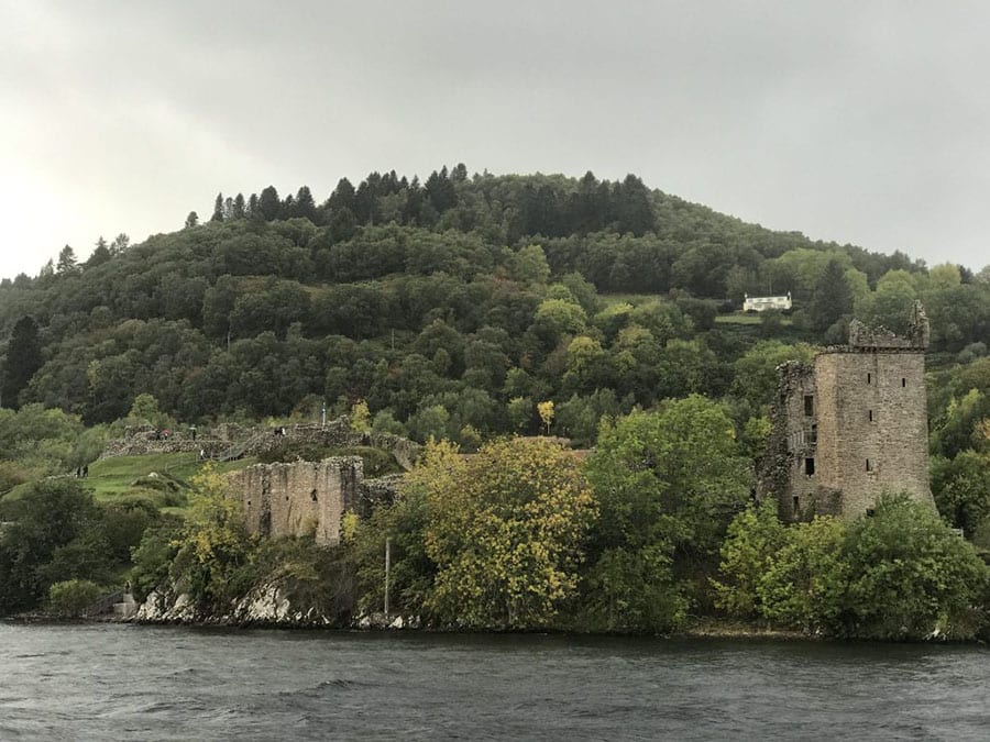 Green wooded hill with the ruins of Urquhart Castle at the bottom on the banks of the Loch Ness