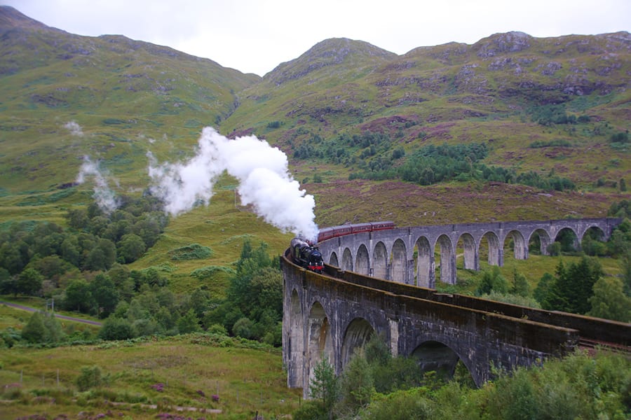 Black and red steam train on a curved viaduct with a series of arches below, green hills in the background
