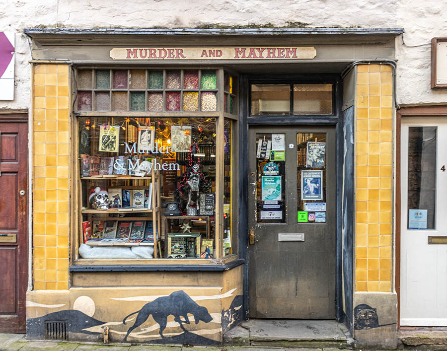 Front of bookshop called Murder and Mayhem with yellow tiles each side of the door, a mural of a hunting dog and a cat under the window.