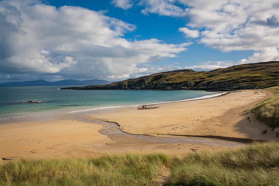 sandy beach with blue sea and green grass to the bottom and hills in the background