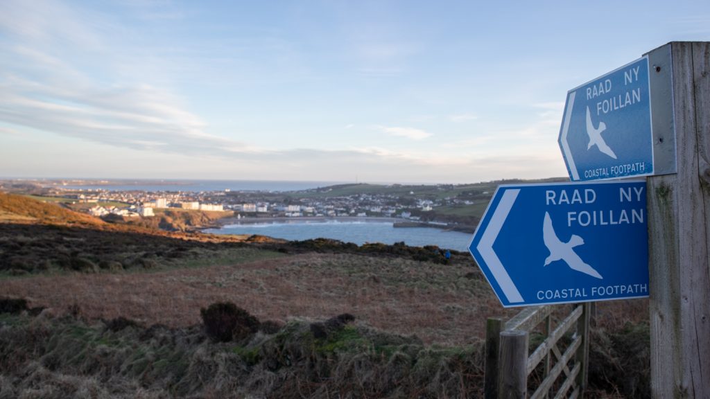 two blue signs saying Raad Ny Foillan with a bay in the background
