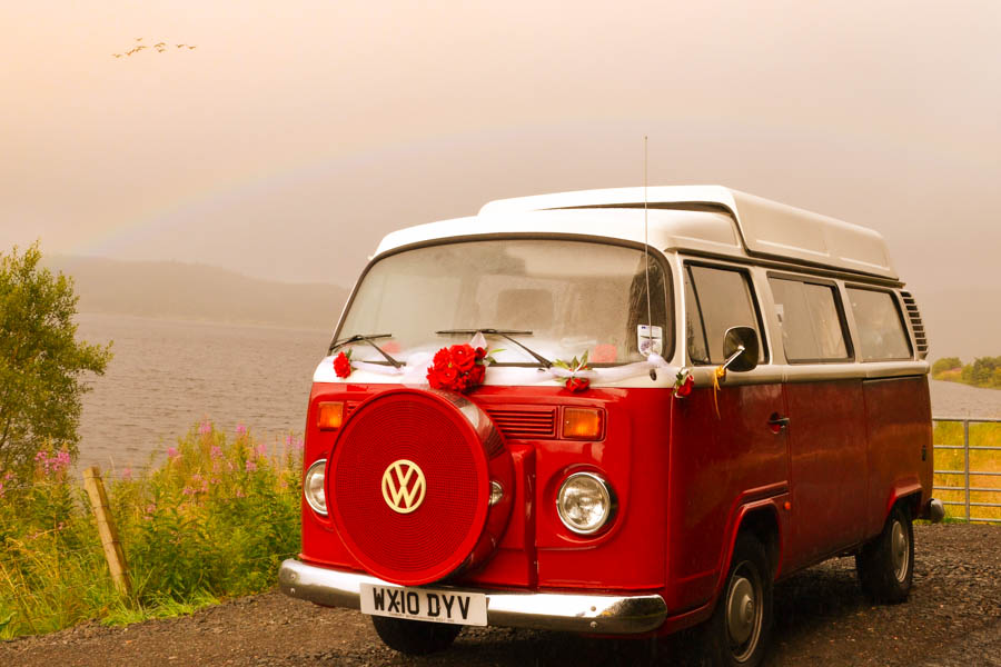 red vw camper vans with flower on the front and a loch in the background with a rainbow above