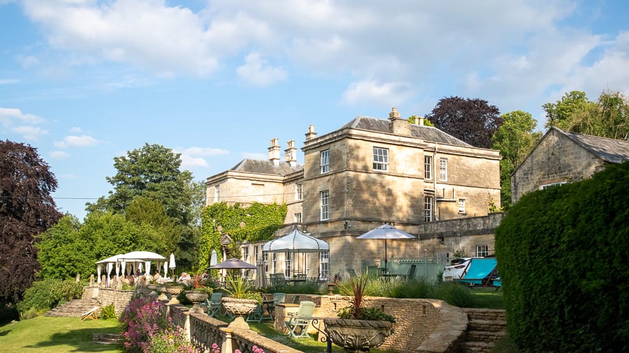 Exterior of the Burleigh Court Hotel, double fronted three storey hotel in creamy Cotswold stone with trees behind and grass in front