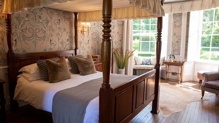 dark wood four-poster bed with mole coloured throw, caramel velvet cushions and pastel pink wallpaper with crested crane bird print in black, white and grey