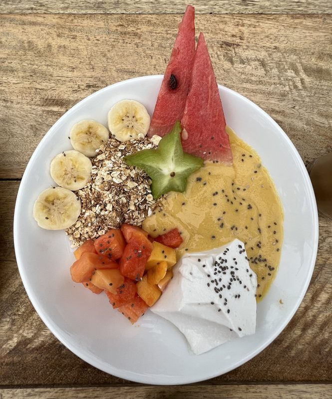 white bowl filled with sliced bananas and starfruit, diced guava, triangles of watermelon, muesli, thick fruit smoothie and vegan coconut yoghurt