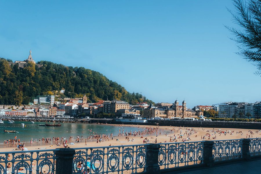 ornate railings above a sandy beach with people on the sand and in the sea and the city of San Sebastian behind