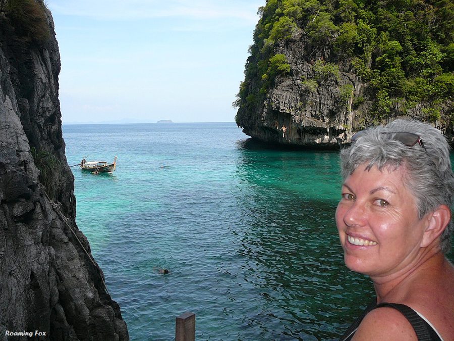 Alma from Roaming Fox on PhiPhi Island, Thailand smiling at the camera with turquoise sea and green covered rocks behind her