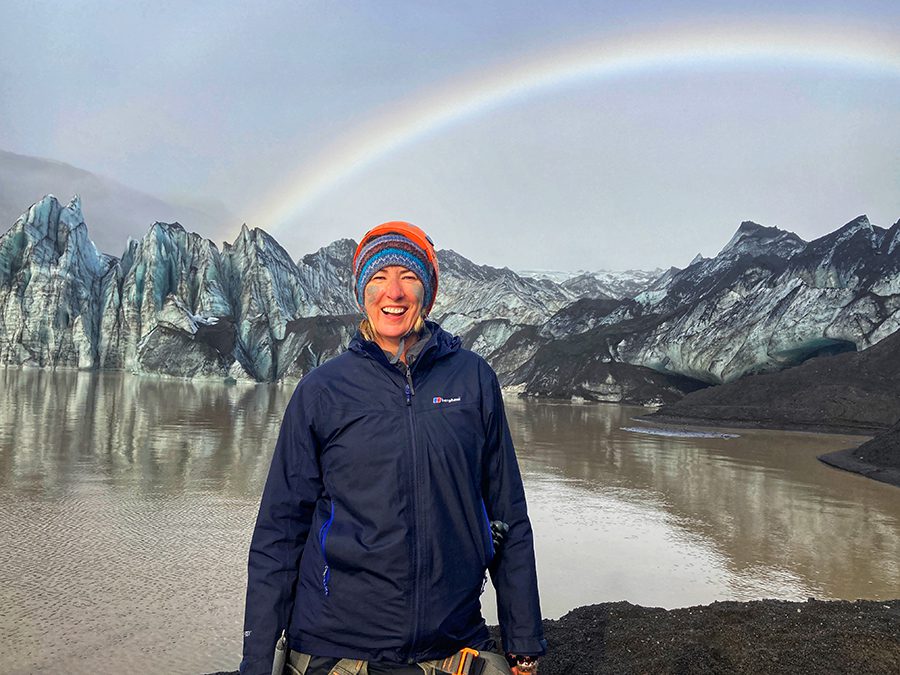 Sue from SueWhereWhyWhat in Iceland wearing a blue jacket and an orange and turquoise woollen hat with mud on her cheeks,with icy cliffs behind her and a rainbow in the sky