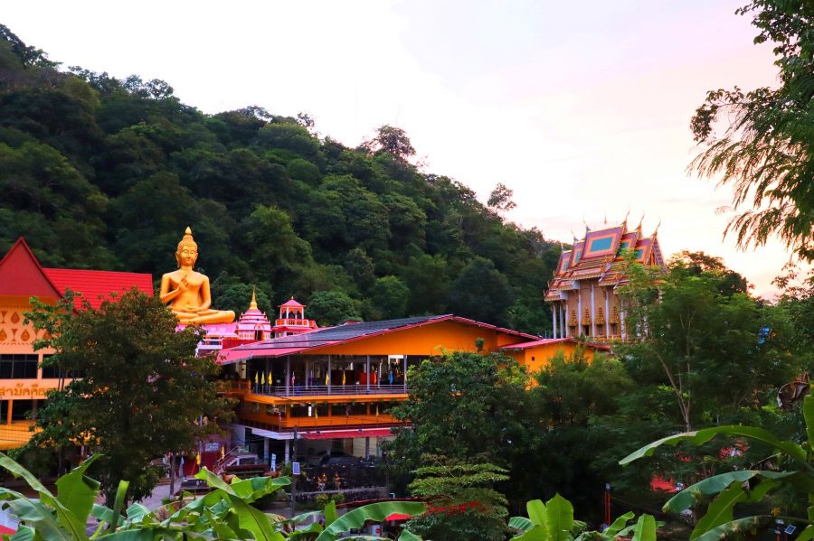 a large golden buddha next to brightly painted buildings in pinks, purples oranges and reds in amongst green trees