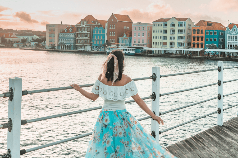 pastel townhouses on the edge of the waters with a woman with her back to the camera wearing a white off-the-shoulder lacy top and turquoise floating floral skirt and a peach scarf in her hair