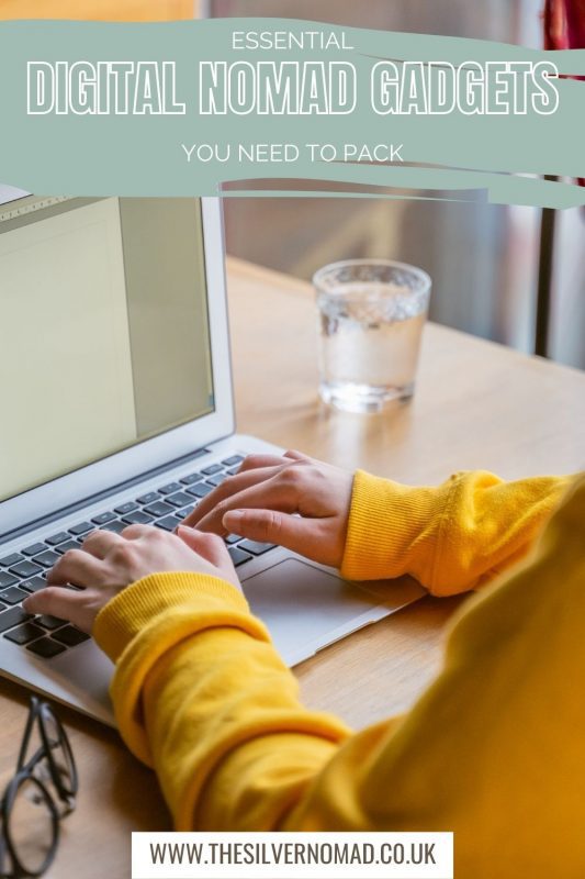 Image of a laptop with someone wearing a yellow sweatshirt typing. Black rimed glasses are to the side and a glass of water on the other side. The words "Essential Digital Nomad Gadgets you need to pack" in capitals on the top