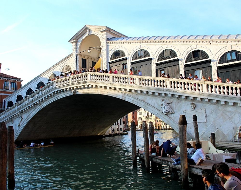 The white Rialton Bridge in Venice with arched doors and people on the bridge as a gondola goes underneath