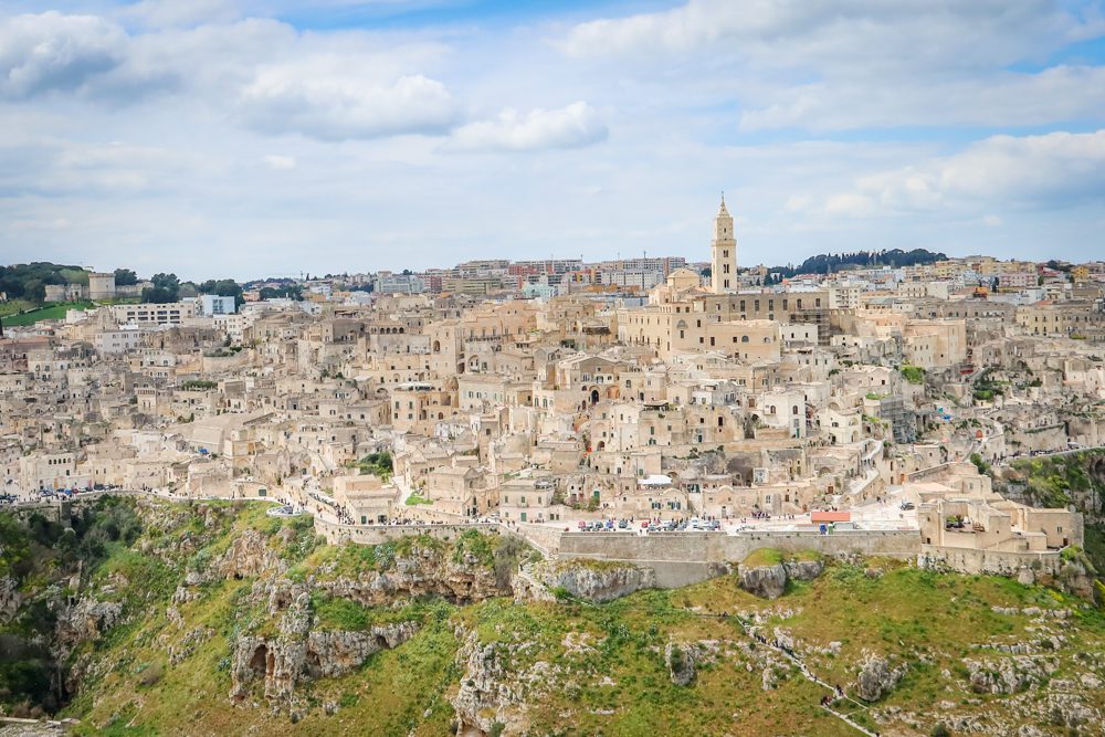 the cream coloured walled city of Matera with church tower prominent. The city is on a hill and the crags are below