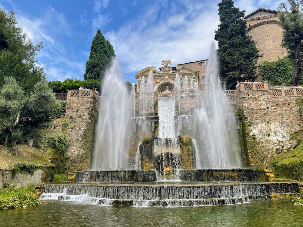 Fountain at Villa d'Este with top level fountains, a waterfall and two main fountains either side flowing into pools below. Behind are cream coloured buildings and green cypress trees