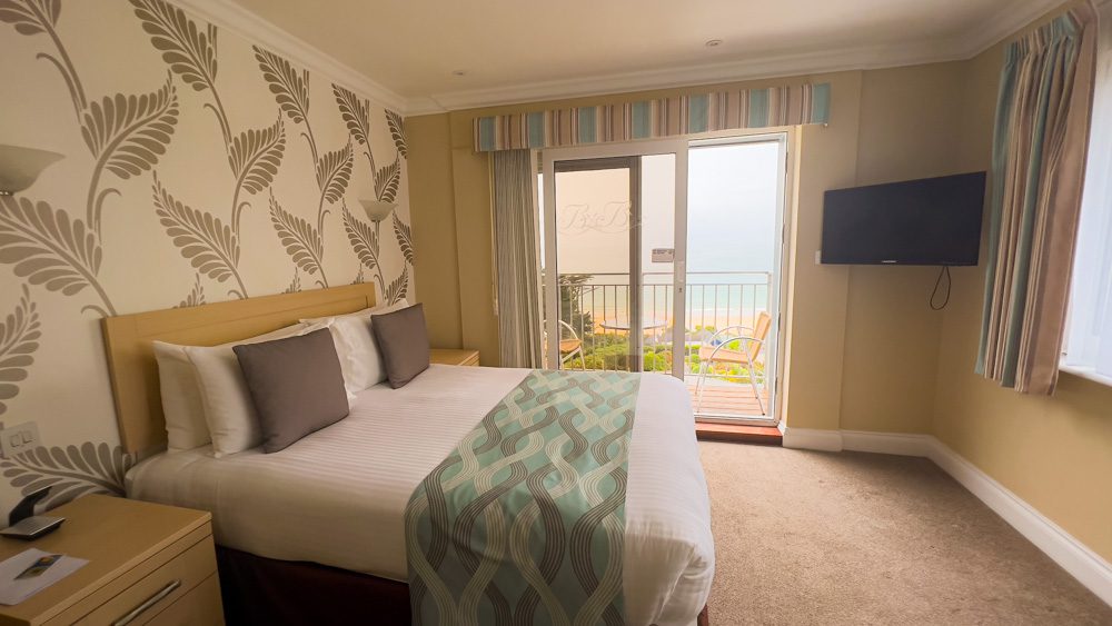 The bedroom at the Biarritz Hotel in Jersey with beige and cream leaf wall paper, pale yellow walls and king size bed with white linen, chocolate coloured cushions and blue, brown and white blanket over the bed. There is a TV screen on the wall in the corner and open patio windows showing a chair and table and the sea view beyond