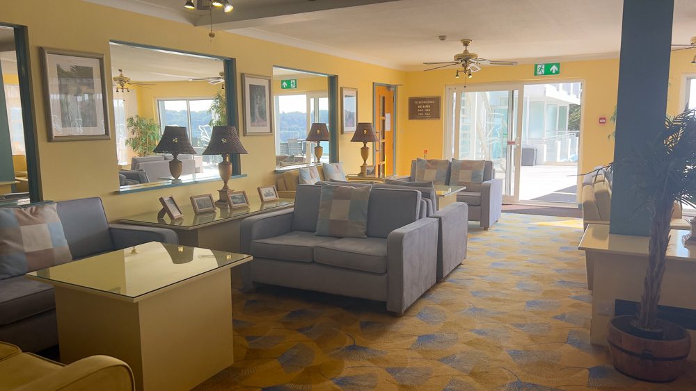 One of the lounges in the Biarritz Hotel with blue-grey couches, blue and yellow carpet and yellow walls with an open sliding door to the terrace