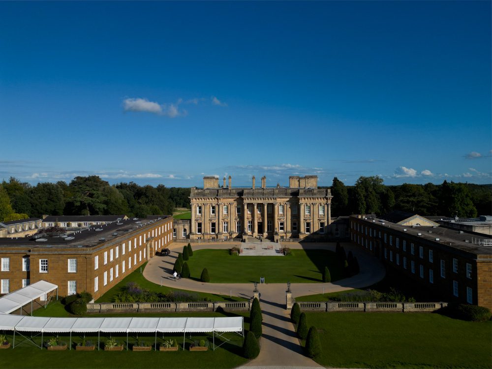 The front of Heythrop Park in the Cotswolds, showing a grand golden stones Georgian building with four columns at the front. There are two wings, one on either side and a white covered walkway to the left