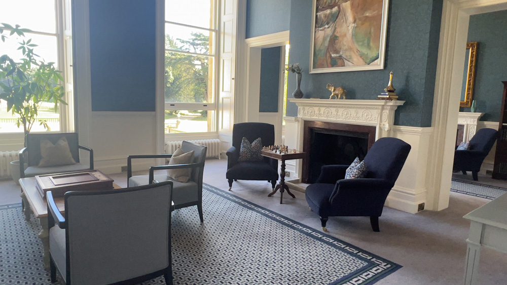 China blue wallpaper with white moulding and 2 navy blue chairs around a chess board. 3 pale blue chairs around a table with a navy and cream geometric rug