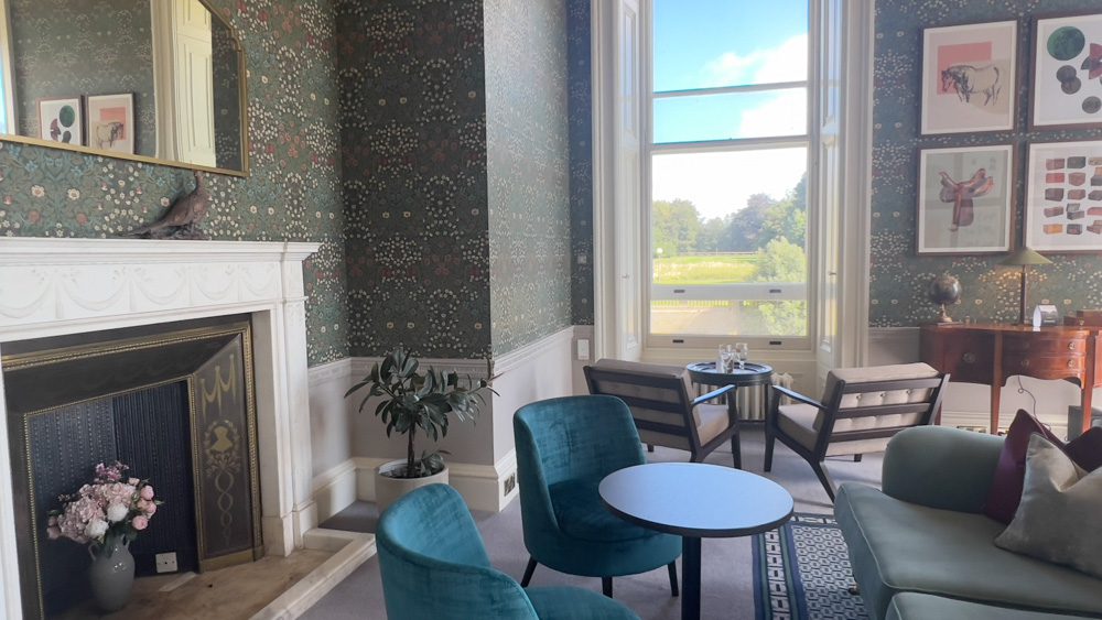 One of the lounges with a fireplace and large mirror. Two blue chairs in front of a sofa with two cream chairs facing out of the window