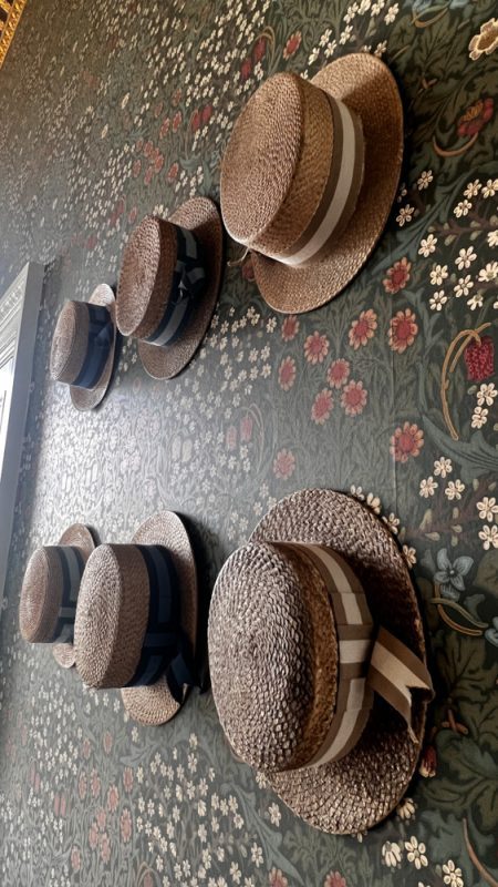 six straw boaters hanging on the wall. The wallpaper is a dark green William Morris design with white and red flowers