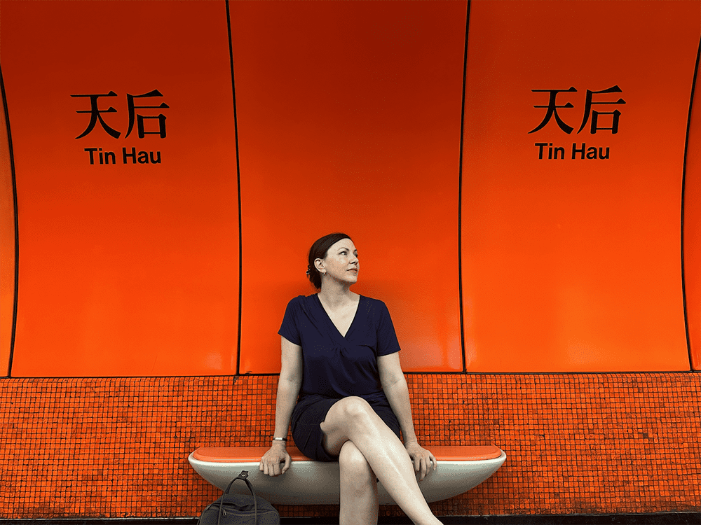 Christa from Beyonderlust in navy short-sleeved dress sitting with her legs crossed on a bench in front of a red wall in a Hong Kong subway with the words Tin Hau and Chinese characters on it

