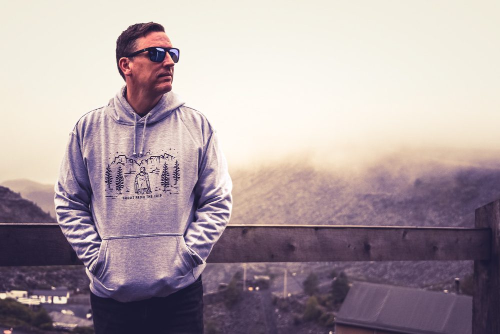 Man standing with his back against a railing wearing a grey hoodie and sunglasses. The hoodie has the words Shoot from the trip in capital letters on it and a design of a man , hills and pine trees
