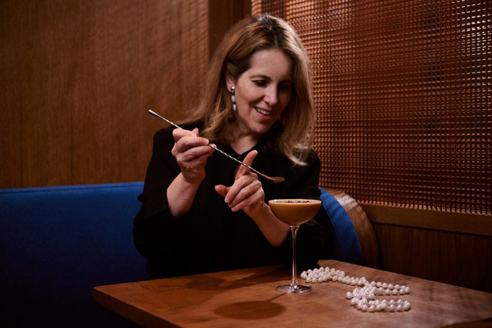 Susan Schwartz. The photo shows a brown haired lady sitting on a blue bench with wicker panel to her right. She is dusting an espresso martini with chocolate using a long silver spoon with a twisted handle. There is a string of pearls on the table. Susan is part of the a more mature bloggers post.