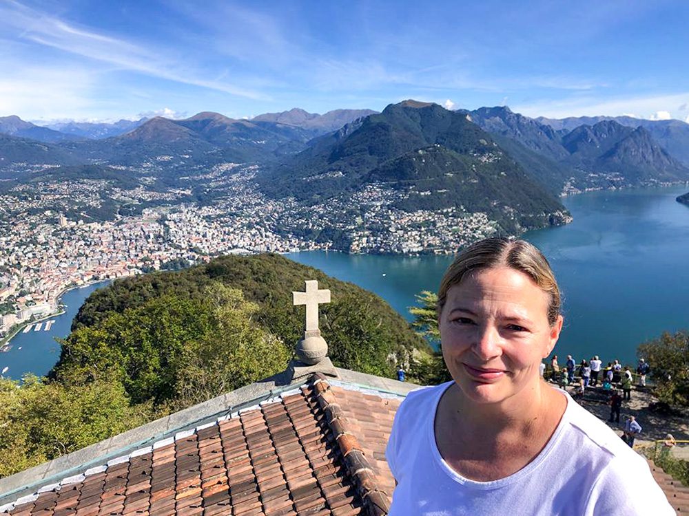 Emma with hair drawn back in to a bun, above Lake Lugano in Switzerland. The far shore is a town with thite houses and red roofs. There are hills in the background and there is a church roof below Emma with a stone cross.