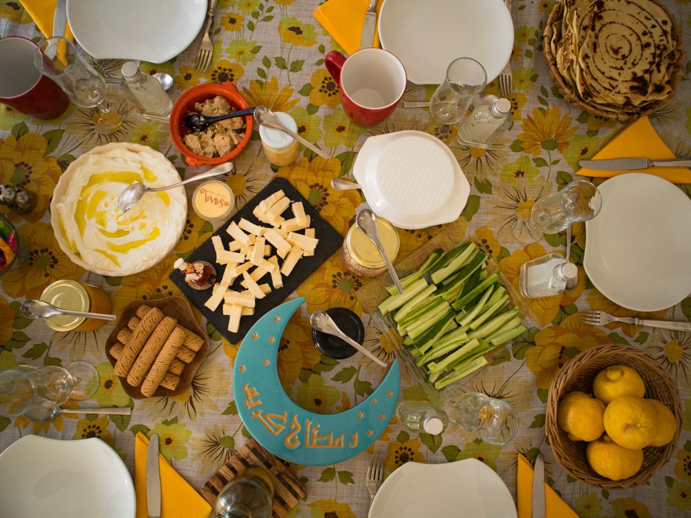 Breakfast table with cut cucumbers, mwali, labneh, halva, a tuquoise crescent moon box with arabic writing on it, white plates, glassed and cups