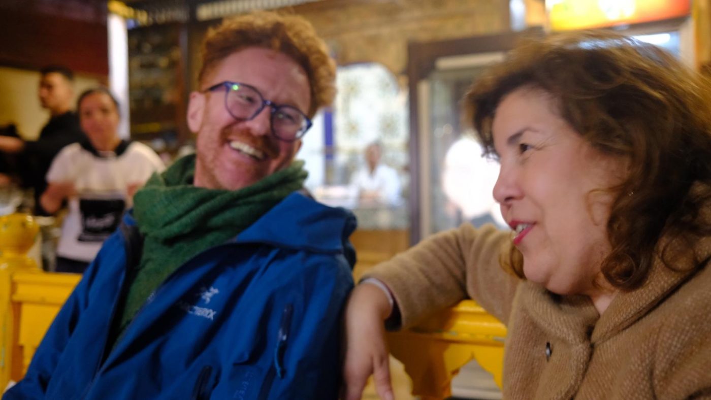 Jamie and Lamia, founders of Sawa Taste of Tunisia. Jamie on the left wearing a blue jacket, green scarf and blue glasses. He has ginger hair and a stubble. Lamia, on the right, wears a brown woolen jacket and had red lipstick. They are sitting on a bright yellow bench which just shows behind them
