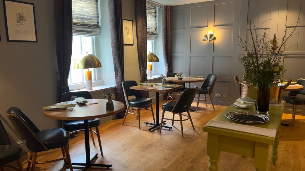 dining room at The Olive Tree Restaurant with soft grey walls, round and square tables with grey leaterher chairs and a lime green console in the middle with a vase of flowers and a silver platter on it.