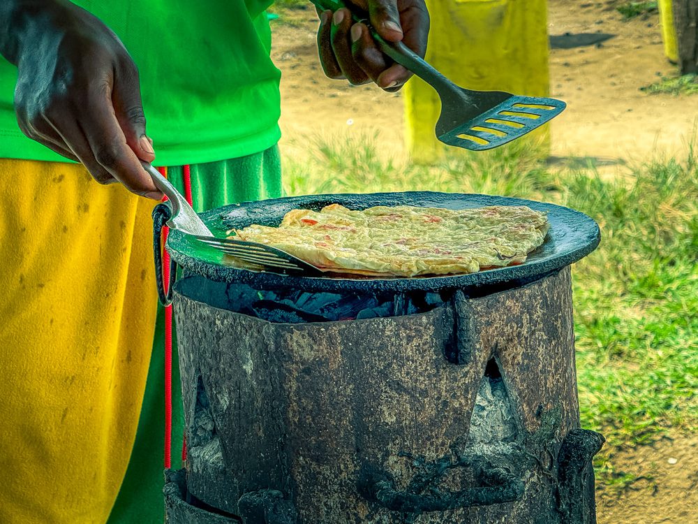 The omelette cooking on the makeshift stove with a man with green top and green and yellow shorts